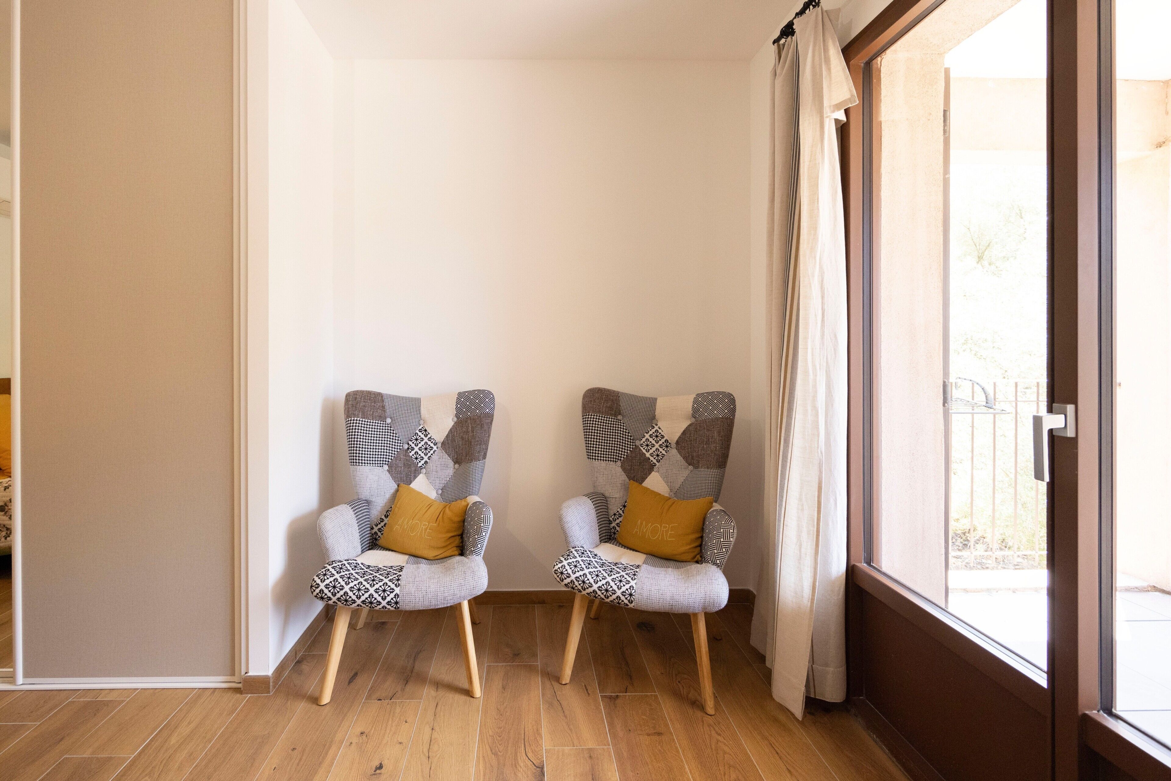 Two armchairs with patterned upholstery and wooden legs, positioned in a corner with a wooden floor and a view through a glass door.