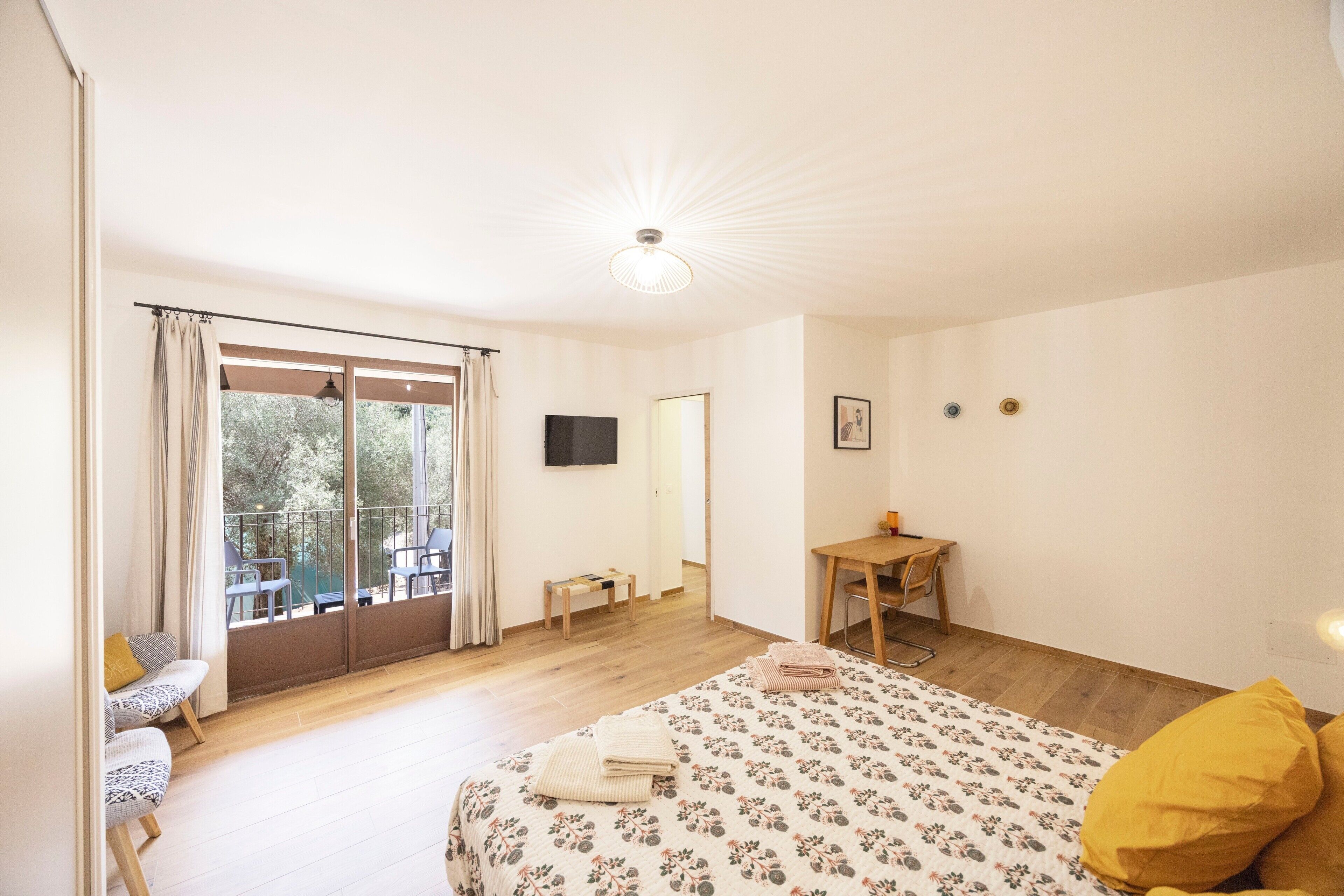 A bedroom with a bed, a desk, a chair, a television, and a balcony with curtains.