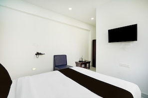 Habitación de hotel con cama, una silla, una mesita con hervidor, televisor de pantalla plana en la pared y toma de corriente.
