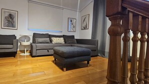 A living room with a grey sofa, a smaller grey armchair, a white side table, and a grey ottoman. There are framed artworks on the walls and a window with blinds.