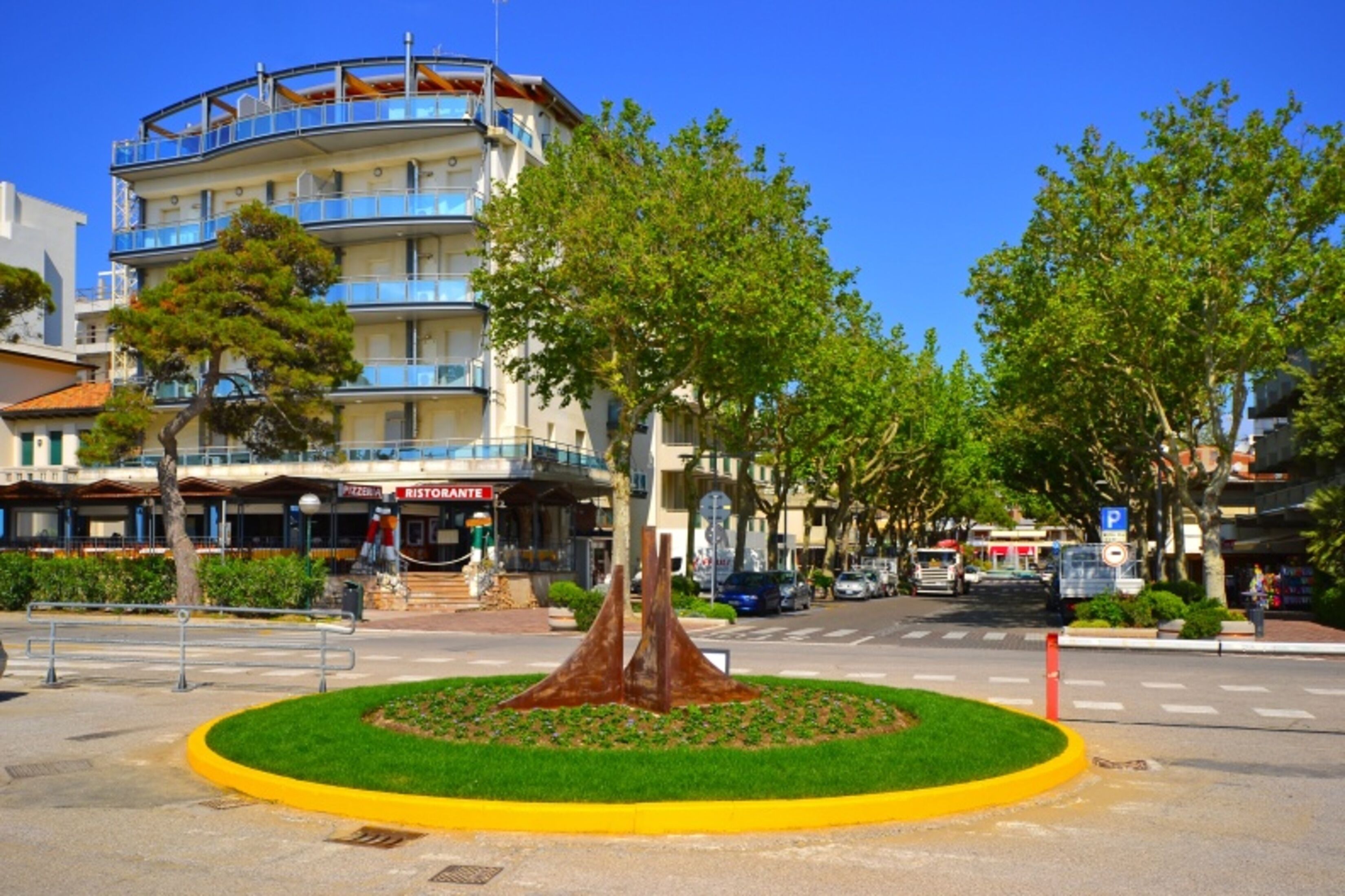 A multi-story building with balconies, a central green area with a sculpture, and a clear blue sky.
