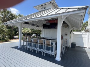 A covered outdoor seating area with metal chairs and a table, surrounded by a white fence and palm trees.