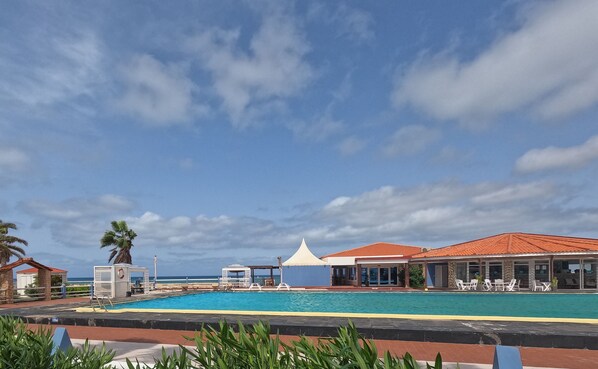 A pool area with lounge chairs, a covered seating area, and a view of the ocean.