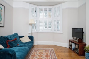 Ein Wohnzimmer mit einem blauen Sofa, einem gemusterten Teppich, einem Fernseher auf einem Holzständer und einem Erkerfenster mit Fensterläden.