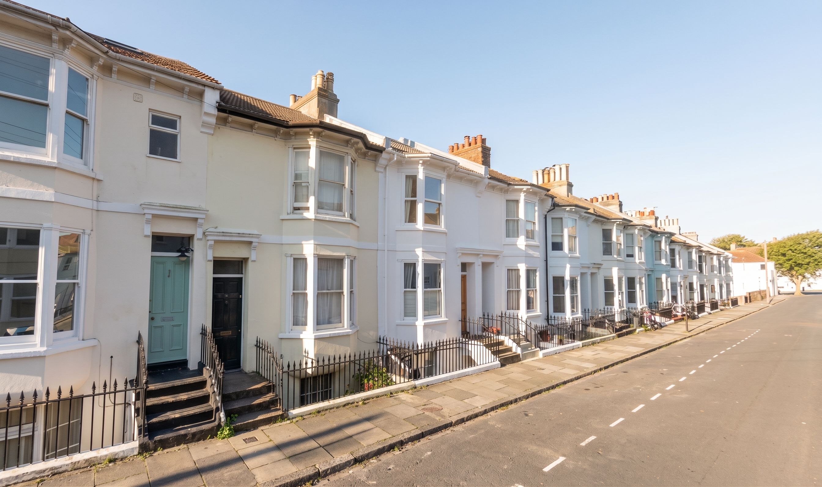 A row of white terraced houses with front doors and windows, a clear sky, and a street.