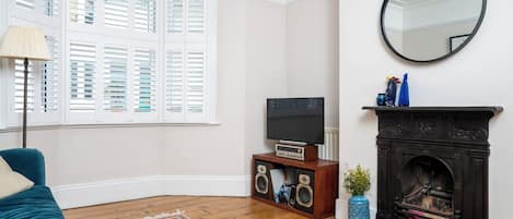 A living room with a television, speakers, a fireplace, a mirror, and a window with shutters.