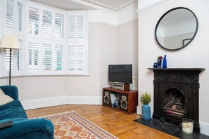A living room with a television, speakers, a fireplace, a mirror, and a window with shutters. - Modern 2BDR Retreat | Near Beach & City Centre (Brighton and Hove)