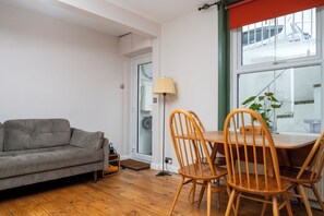 A living room with a grey sofa, wooden chairs, a table, a floor lamp, and a window with a view of a building. - Modern 2BDR Retreat | Near Beach & City Centre (Brighton and Hove)