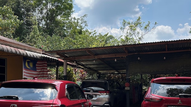A parking area with three cars, a covered structure, and a building with a Malaysian flag.