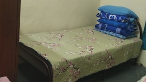 A small room with a single bed, a wooden wardrobe, and a stack of folded blankets.