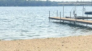 A calm lake with a wooden dock extending into the water, a covered seating area with a fan, and a sandy shore. - Daggett Lake Cabin with Dock & Beach (Crosslake)