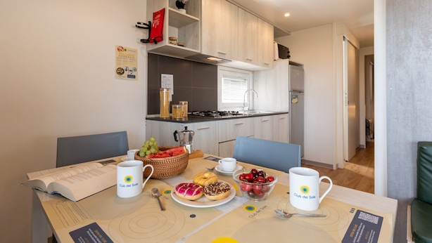 A modern kitchen with a dining table set for breakfast, including fruits, pastries, and coffee mugs. - Jesolo Family Resort (Jesolo)