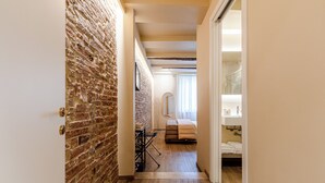 Een smalle gang met een bakstenen muur, een bed met een bruine deken, een kleine tafel met een stoel, en een badkamer met een wastafel en handdoekhouder.