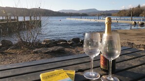 Een houten tafel met twee lege wijnglazen en een champagnefles, een kaart, en uitzicht op een meer met bergen op de achtergrond.