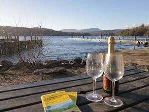 A wooden table with two empty wine glasses and a bottle of champagne, a map, and a view of a lake with mountains in the background. - Wilders Wood (Windermere)