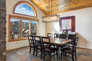 A dining area with a large table and chairs, a hanging light fixture, a window with a view of a forest, and a framed picture on the wall.