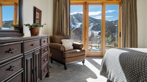 Un dormitorio con un ventanal grande, un aparador de madera, un sillón marrón y vistas a montañas.