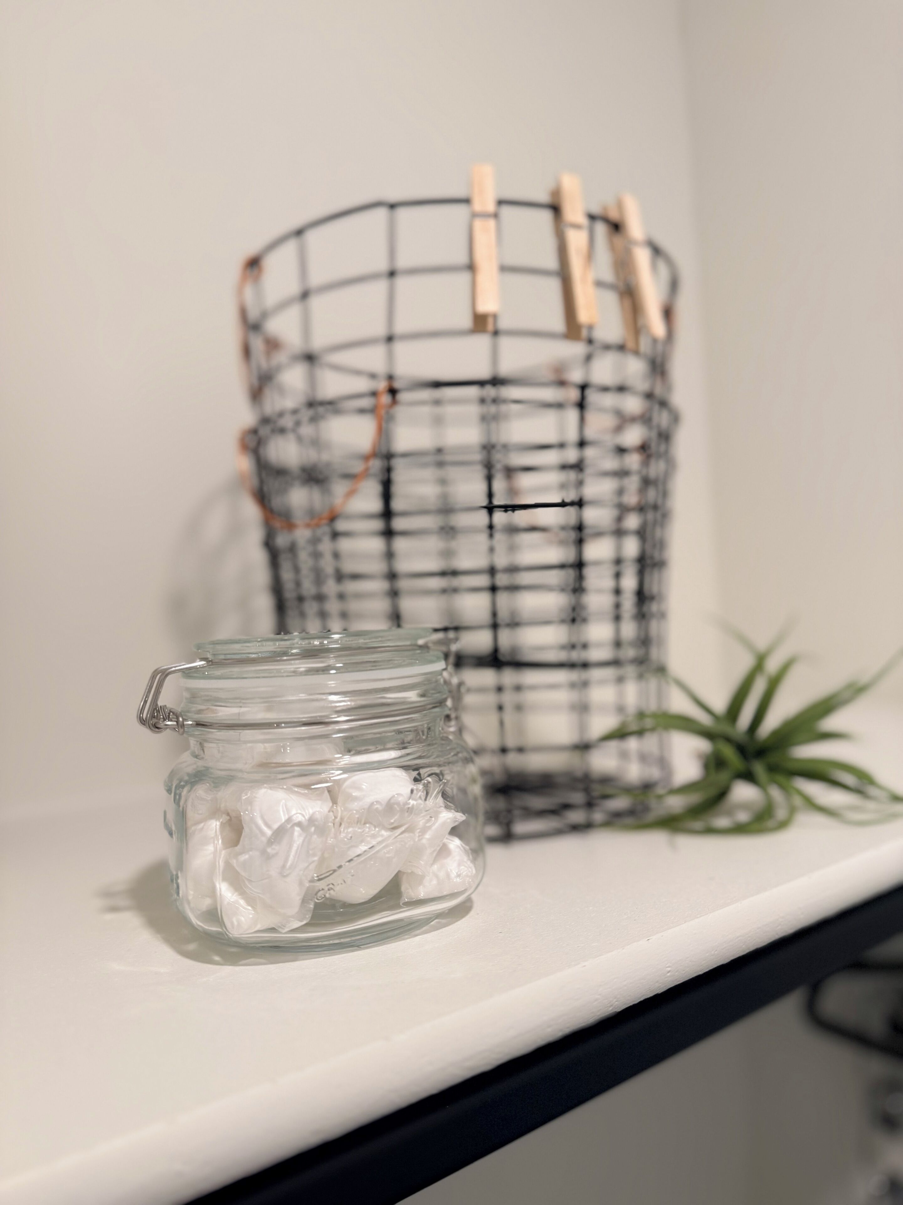 Una estantería con un frasco de vidrio que contiene objetos blancos, una cesta de alambre con pinzas para ropa y una planta verde.