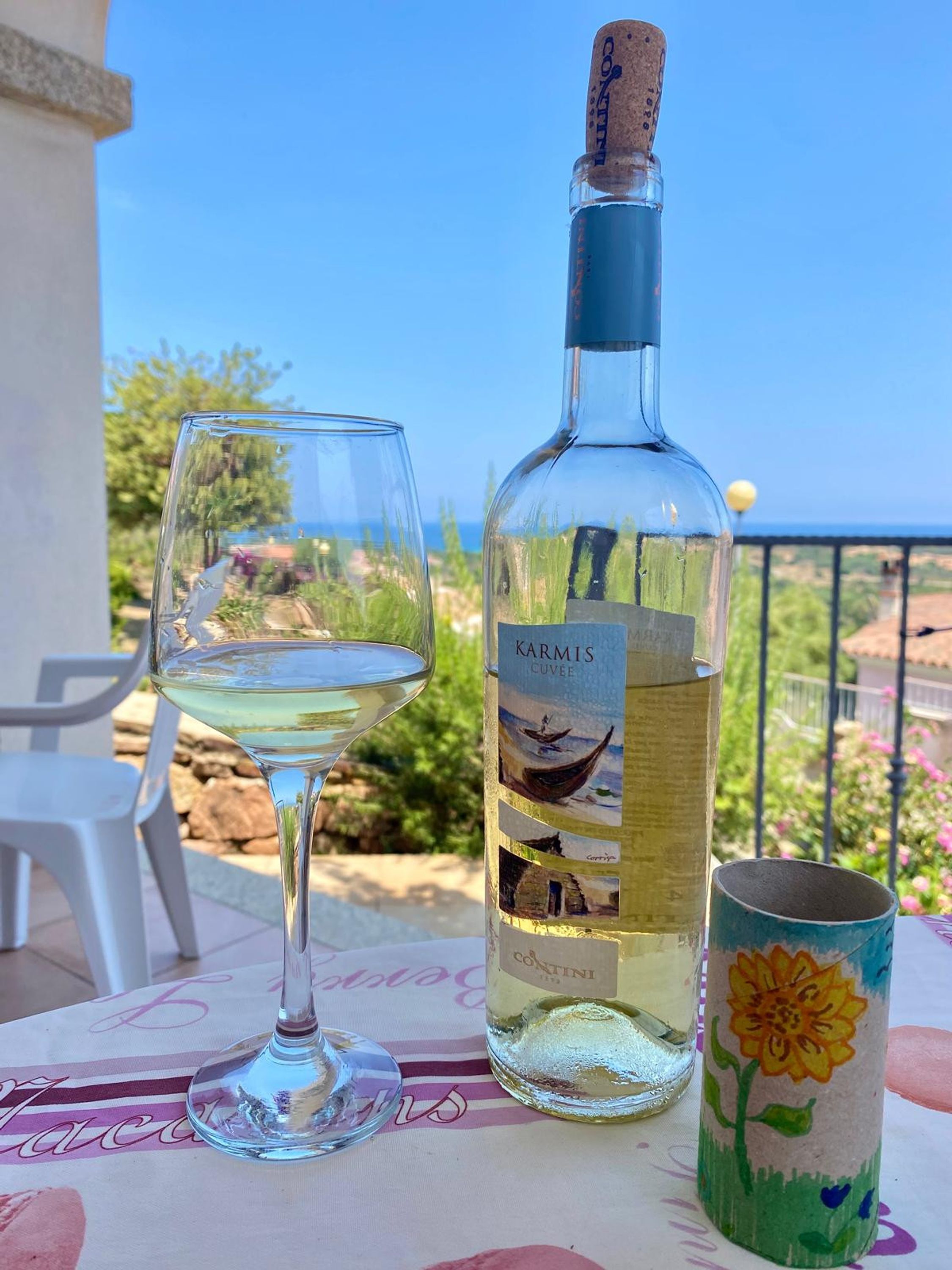 Une bouteille de vin Karmis, un verre de vin blanc et une tasse décorée posés sur une table, avec vue sur la mer.