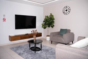 A modern living room with a flat-screen TV, a potted plant, a grey sofa, and a clock on the wall. - Kingston Horizon Retreat Apt (Kingston)