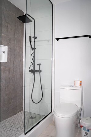 A glass-enclosed shower with a black hose and a handheld showerhead, a white toilet, and a small trash bin. - Kingston Horizon Retreat Apt (Kingston)