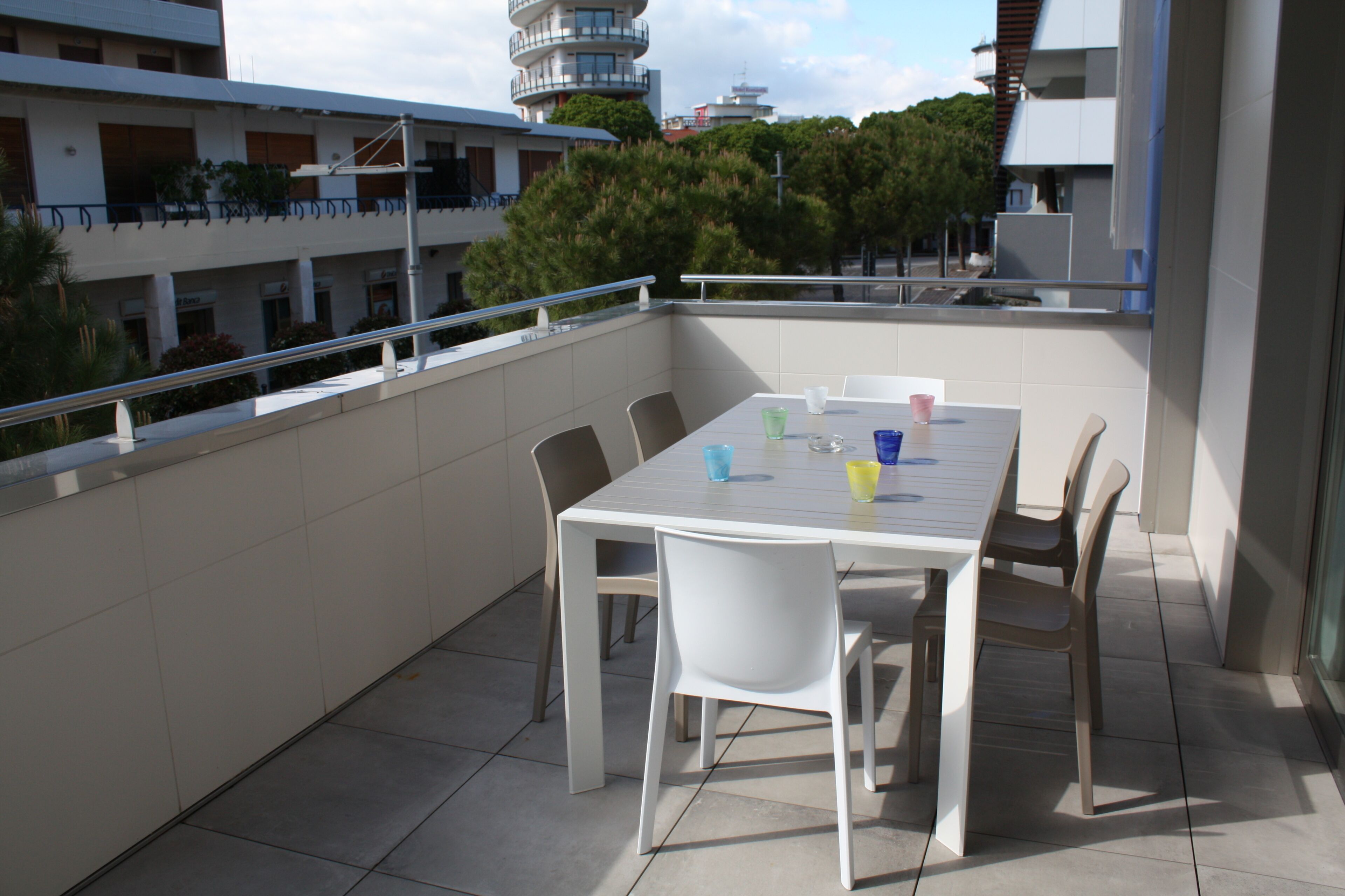 A balcony with a white table and chairs, a railing, and a view of buildings and trees in the background.