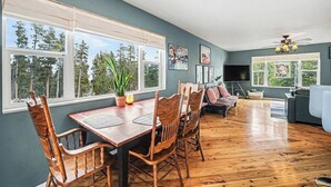 Un coin repas avec une table et des chaises en bois, un canapé et un téléviseur, dans une pièce dotée de grandes fenêtres et d’un ventilateur de plafond.