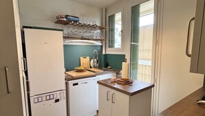 Una cocina compacta con refrigerador, lavavajillas, encimera de madera, ventana con vistas y repisa con libros y vasos.
