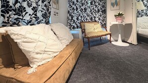 Une chambre à coucher avec un lit, une chaise, une petite table sur laquelle se trouve un vase, et des rideaux à motifs.