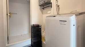 Une chambre d’hôtel compacte avec une porte blanche, un petit chariot de rangement noir, un lave-linge blanc et un porte-serviettes avec des serviettes pliées.