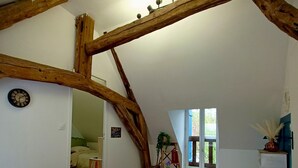 A bedroom with a sloped ceiling, wooden beams, a bed with floral bedding, a small table, and a window with a view. - Gite "La Greniette (Châteauneuf-sur-Loire)
