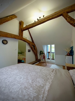 A bedroom with a sloped ceiling, wooden beams, a bed with floral bedding, a small table, and a window with a view. - Gite "La Greniette (Châteauneuf-sur-Loire)