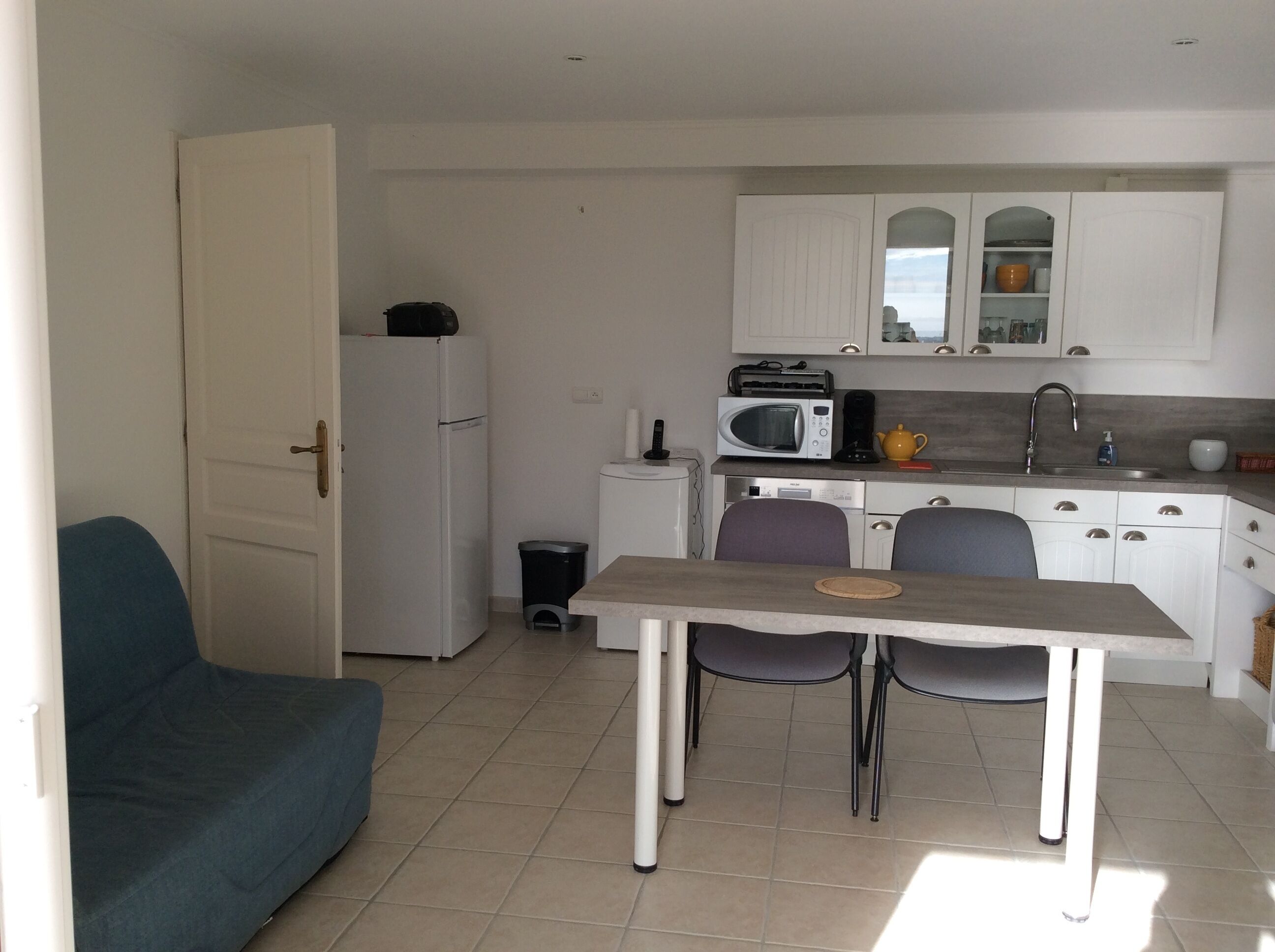 A compact kitchen with white cabinets, a microwave, a toaster, and a sink. There is a small trash bin and a blue chair.