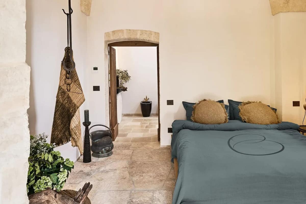 A bedroom with a bed, a nightstand, a lamp, a hanging lamp, a plant, and a stone wall.