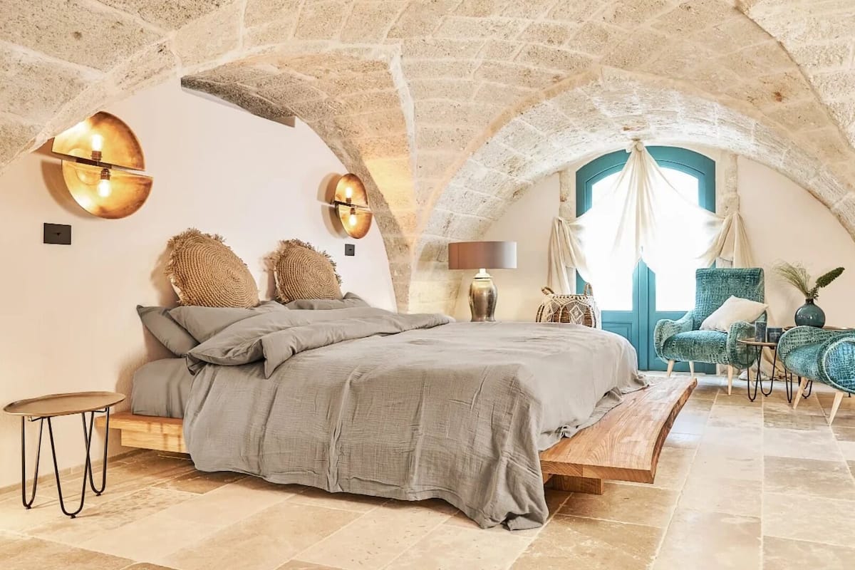 A bedroom with a stone ceiling, a bed with grey bedding, a nightstand, a lamp, and a chair.