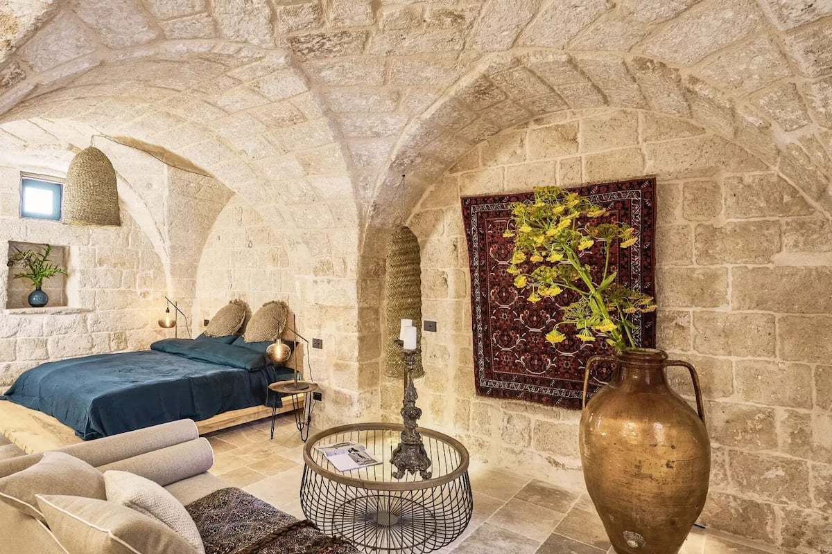 A stone-walled room with a bed, a hanging light fixture, a vase with flowers, and a patterned tapestry.