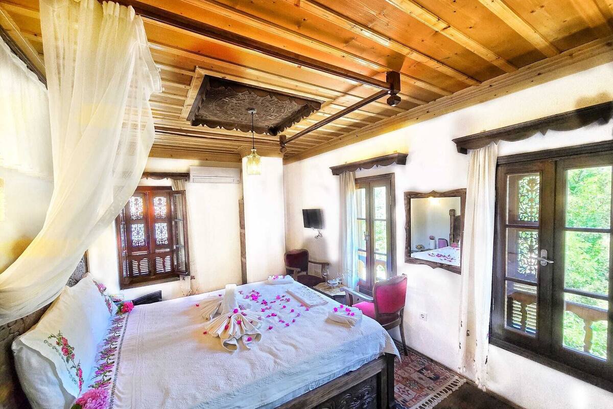 A room with a bed, a chair, a table with a mirror, and a window with a view of greenery.