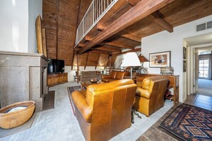 Ein geräumiges Wohnzimmer mit hoher Decke, Holzbalken, Ledersofas, einem Kamin und einem Fernseher.