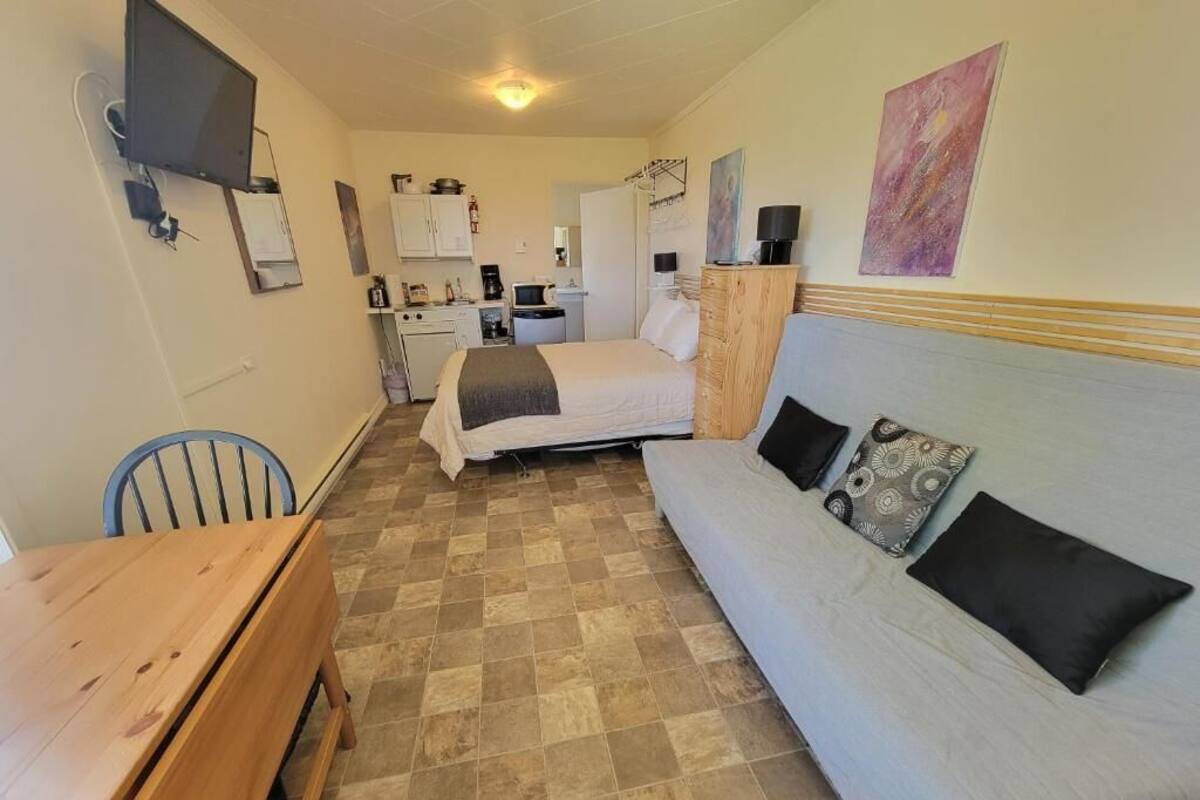 Una habitación compacta con cama, sofá, mesa de madera, televisor montado en la pared y una kitchenette al fondo.