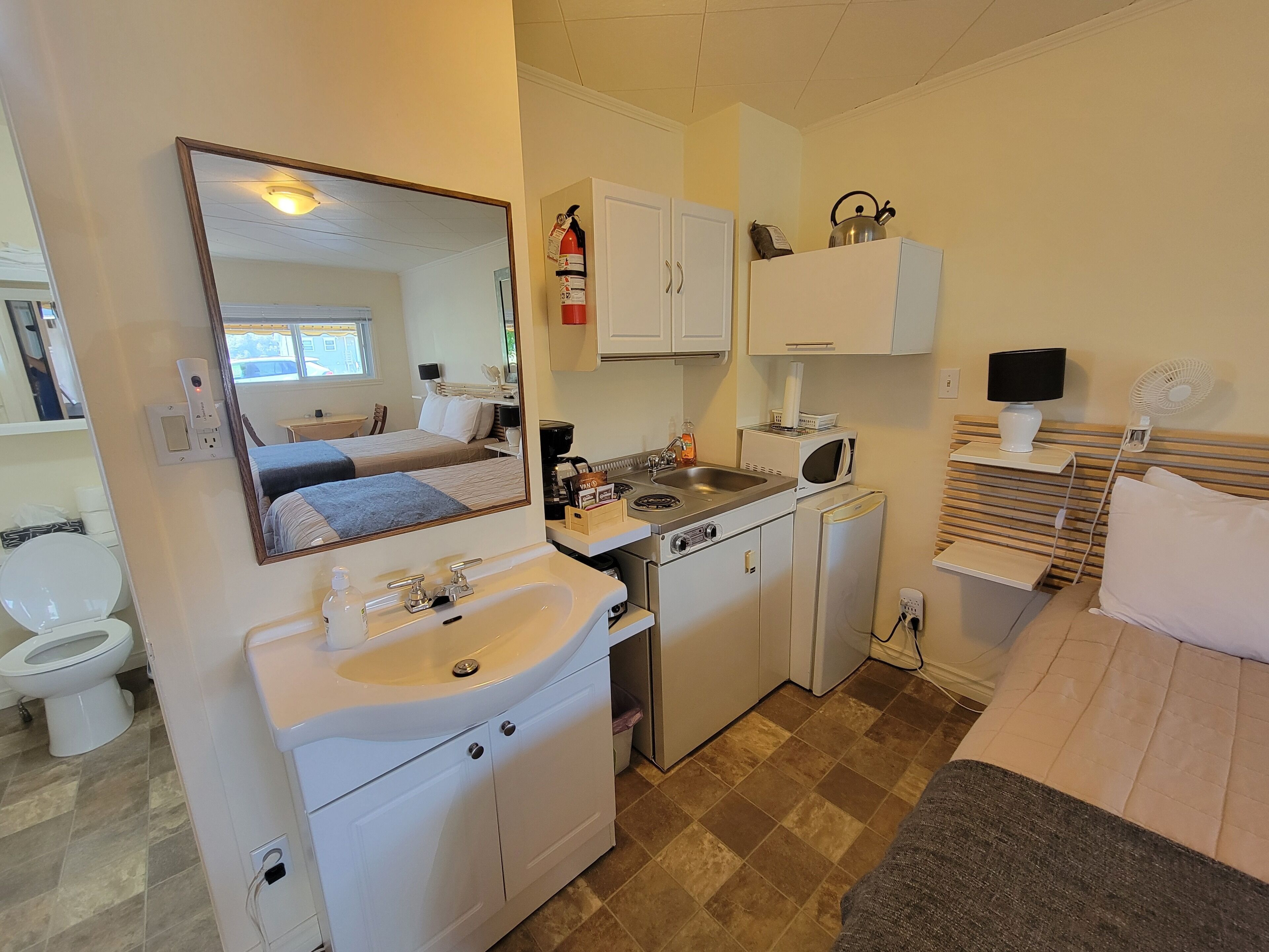 Habitación de hotel pequeña con cama, kitchenette, baño con inodoro y espejo que refleja la habitación.