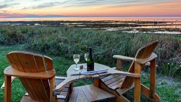 Een houten tafel met stoelen buiten, met een fles en glazen erop, met uitzicht op een waterpartij en een gazon.