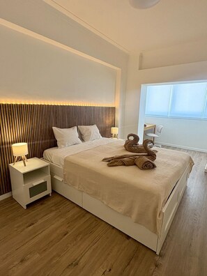A modern bedroom with a large bed, bedside tables, a headboard with vertical lines, and a window with blinds. - Bright Tejo View Apartment - Walk to Ferry - Peaceful Cacilhas Stay (Almada)