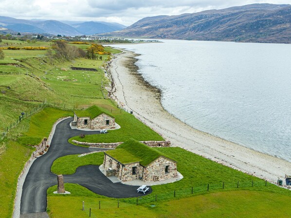 Une route côtière bordée de bâtiments en pierre, avec une zone herbeuse et une vue sur la mer et les montagnes.