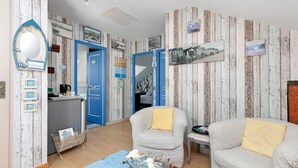 Una habitación con pared de madera, una puerta azul, dos sofás blancos con cojines amarillos, una mesa de centro de vidrio con flores y una mesita con un libro.