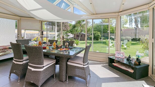 Un comedor con mobiliario de mimbre, una mesa de vidrio y vistas a un jardín a través de amplios ventanales.