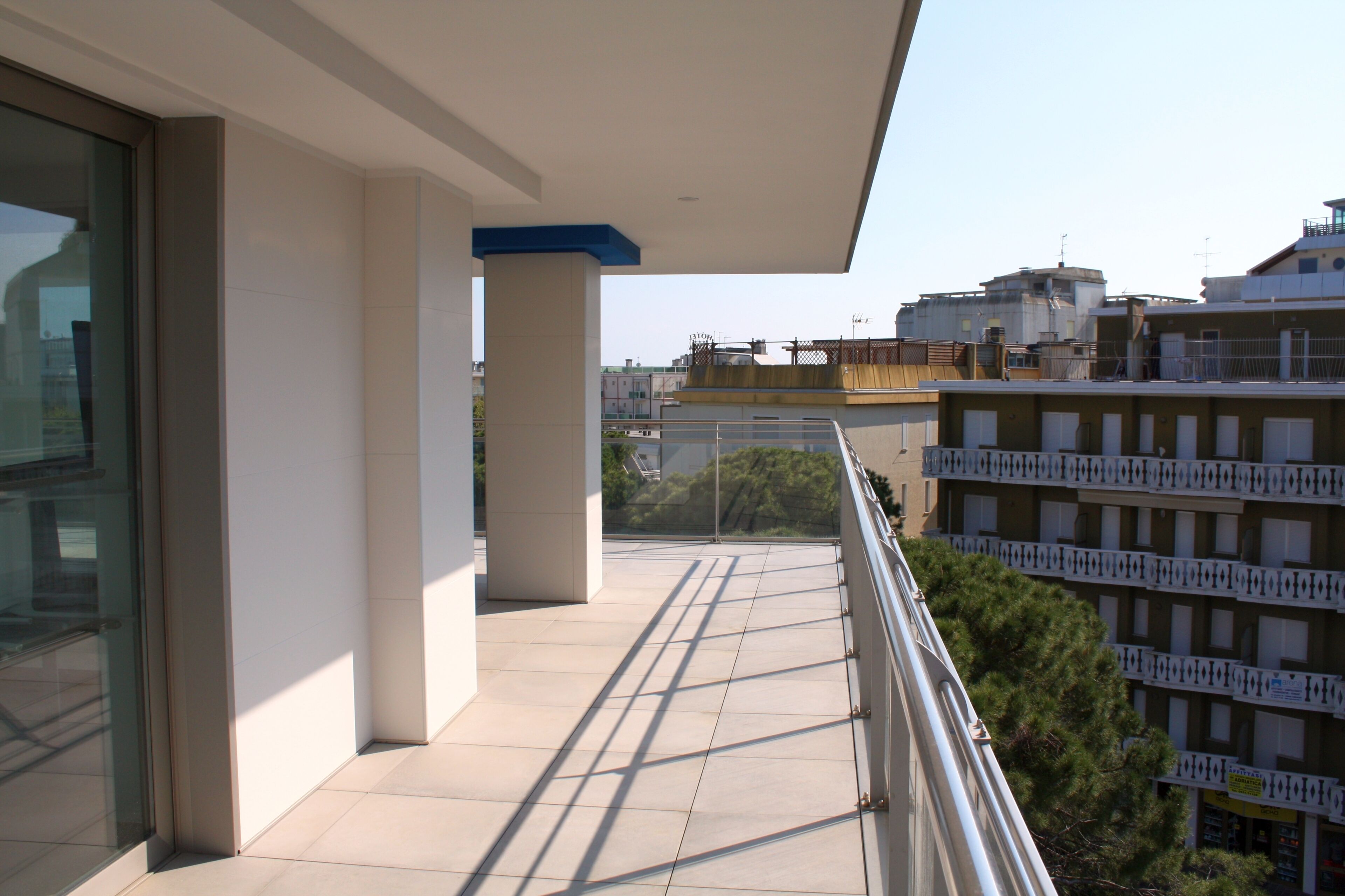 A balcony with a tiled floor and a glass door, overlooking a cityscape with buildings and trees.