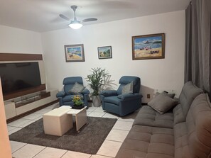 A living room with a flat-screen TV, two blue armchairs, a beige ottoman, a large sofa, a ceiling fan, and two framed paintings on the wall. - Vacation home with a pool, just 150 meters from the beach in Piratininga (Niterói)