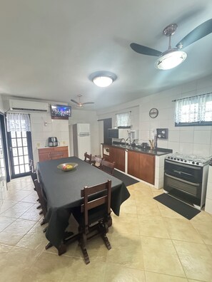 A dining area with a long table, chairs, a ceiling fan, a TV, and a kitchen with a stove and oven. - Vacation home with a pool, 150 m from the beach in Piratininga (Niterói)
