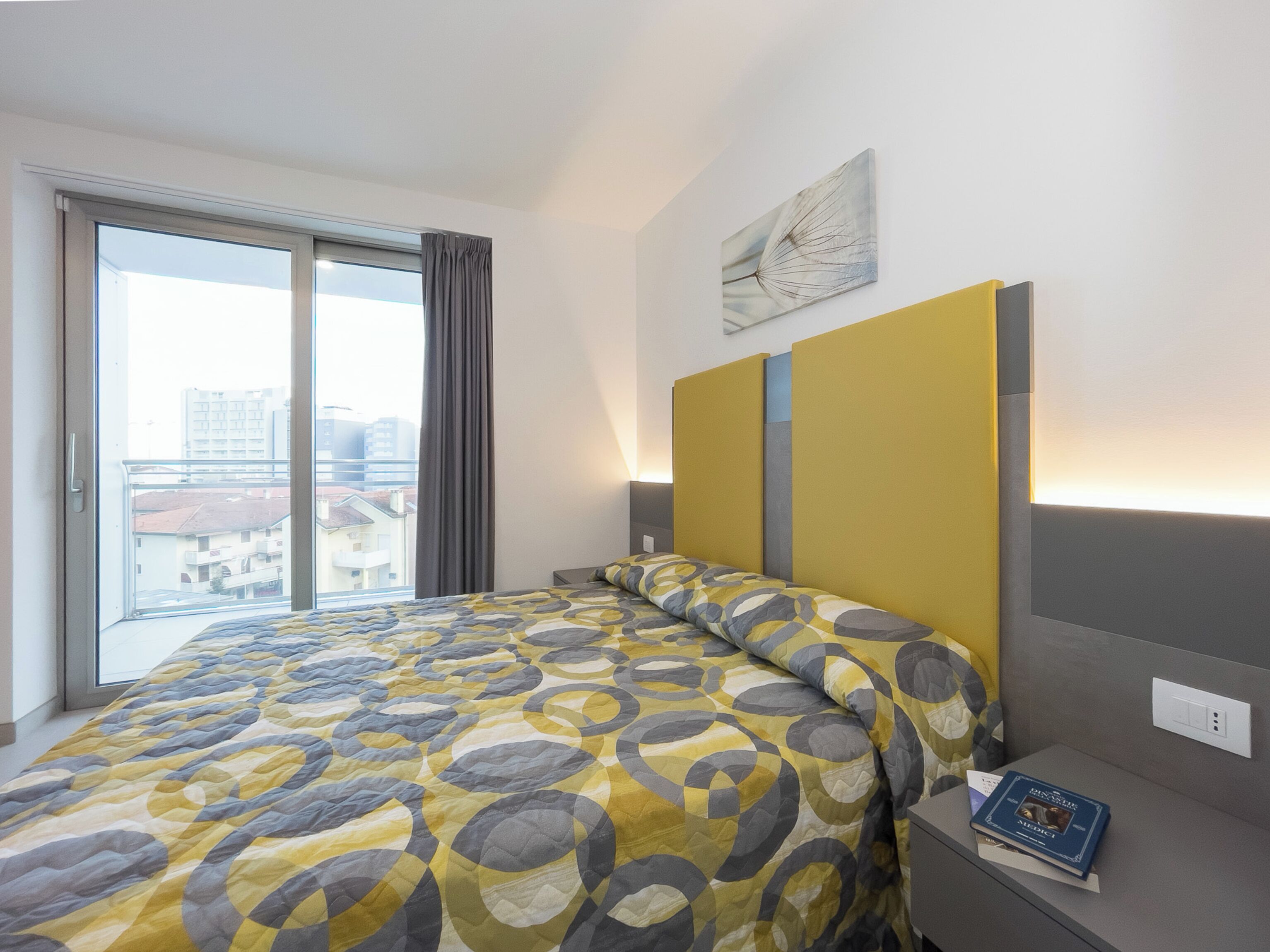 A hotel room with a large bed, a view of the city through a window, a bedside table with a book, and a wall-mounted light fixture.