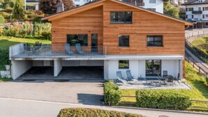 Une maison moderne à deux étages, dotée d’une façade en bois, d’un balcon avec des sièges et d’une allée menant à l’entrée.